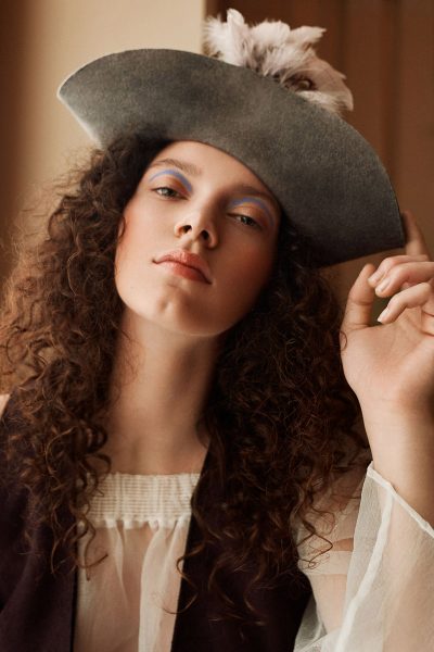 Junge Frau mit Locken, trägt Hut im Piratenstil, blauer Lidschatten als Make-up Inspiration