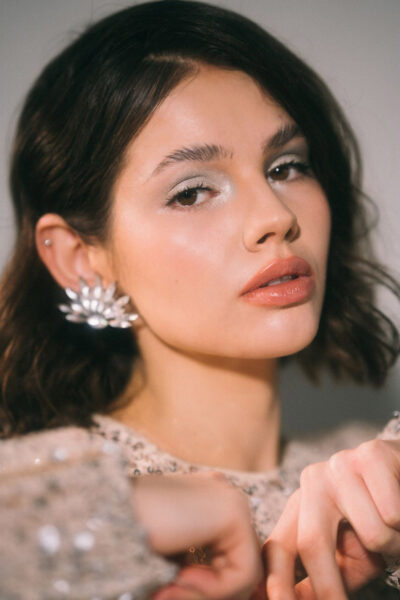 Portrait brünettes Model mit silberfarbenem Cream Eyeshadow, mit Browsoap hochgekämmten Augenbrauen, einem lockigen Longbob mit Zickzackscheitel blickt in die Kamera & hält ihre Hände aneinander